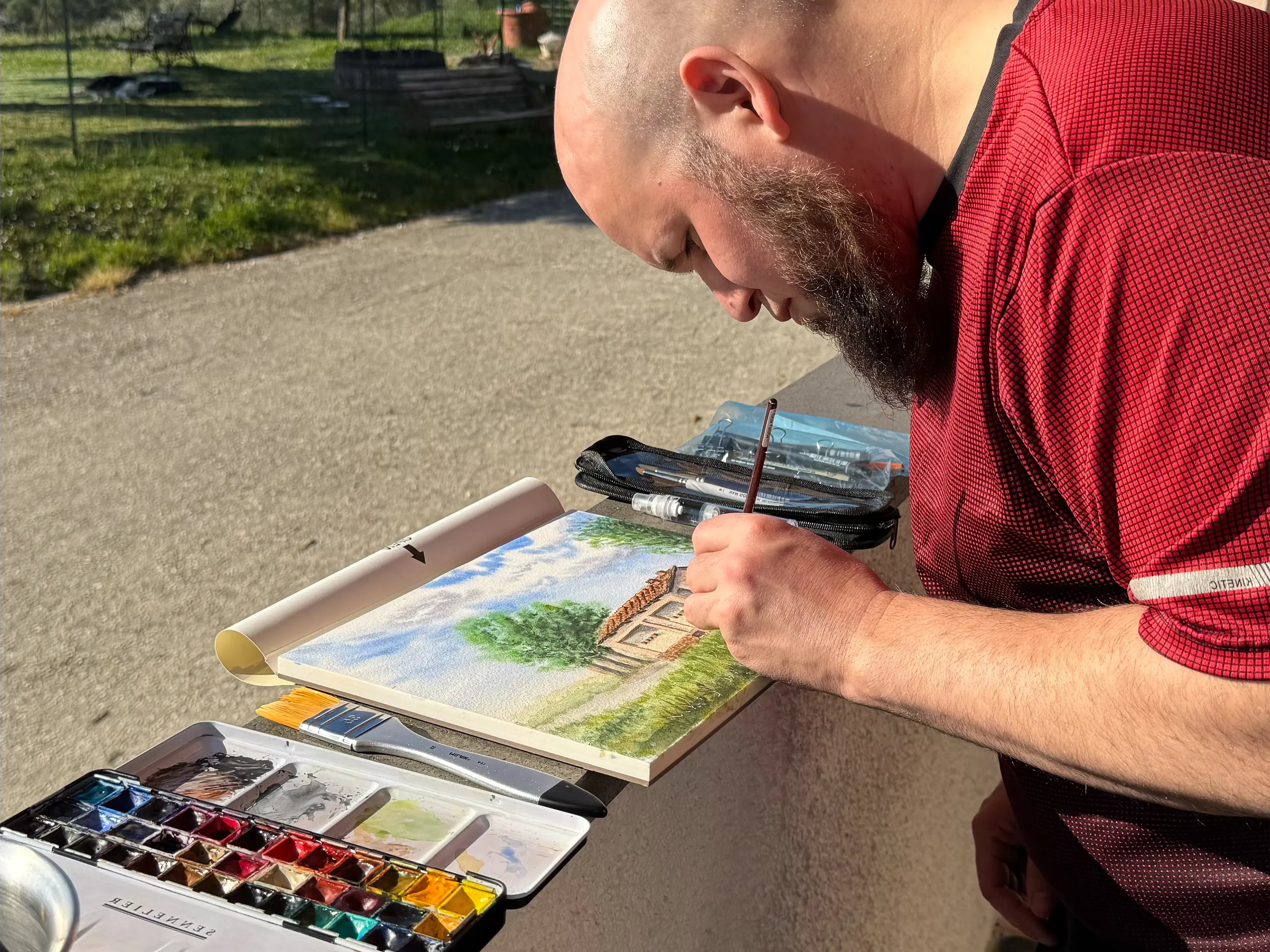 Teobaldo Corro pintando una acuarela al aire libre, inclinado sobre un cuaderno con una paleta de acuarelas y pinceles junto a él, con césped y árboles al fondo.