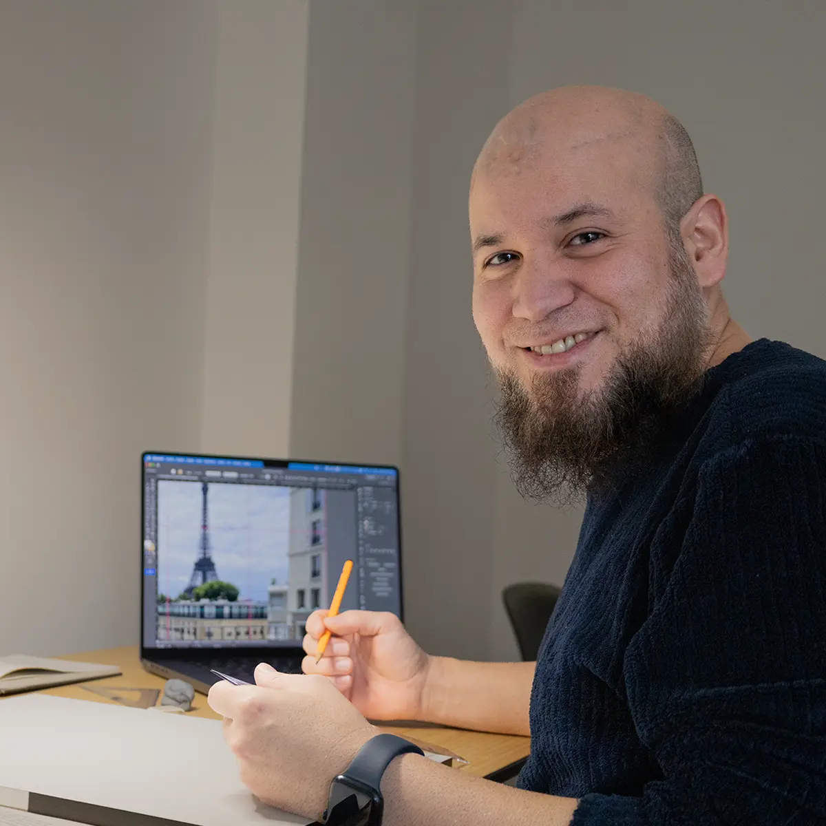 Teobaldo Corro sonríe sentado a su mesa de trabajo, lápiz en mano, junto a un portátil con una fotografía de referencia de la Torre Eiffel abierta en un programa de diseño.